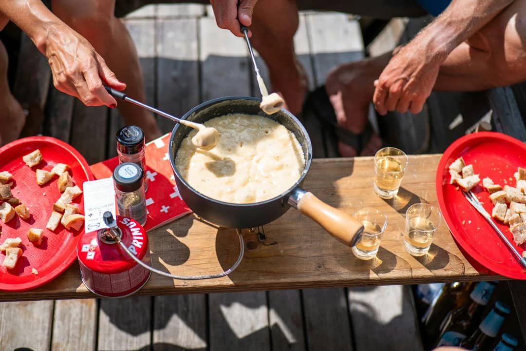 Des amis trempent des dés de pain dans une fondue au fromage partagée, sur une table de terrasse en bois