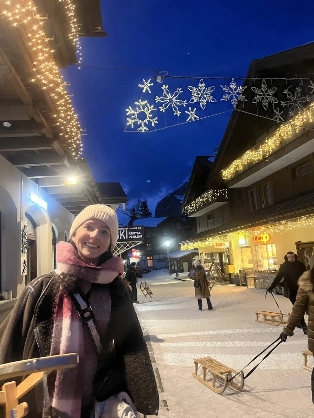 Ein Gast lächelt nachts in einer Strasse in Mürren, während ein Schlittler einen Holzschlitten unter der Weihnachtsbeleuchtung zieht