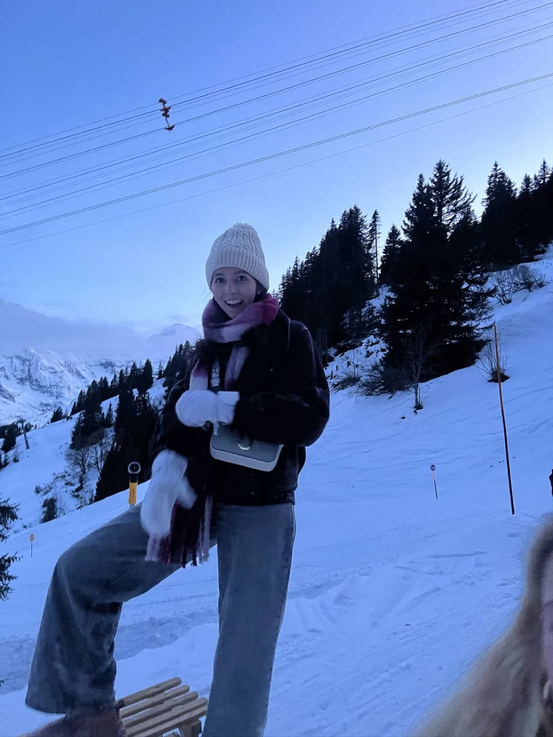 Gast mit rosa Mütze stellt einen Schuh auf einen Holzschlitten an einem verschneiten Schlittelhang oberhalb von Mürren in der Dämmerung