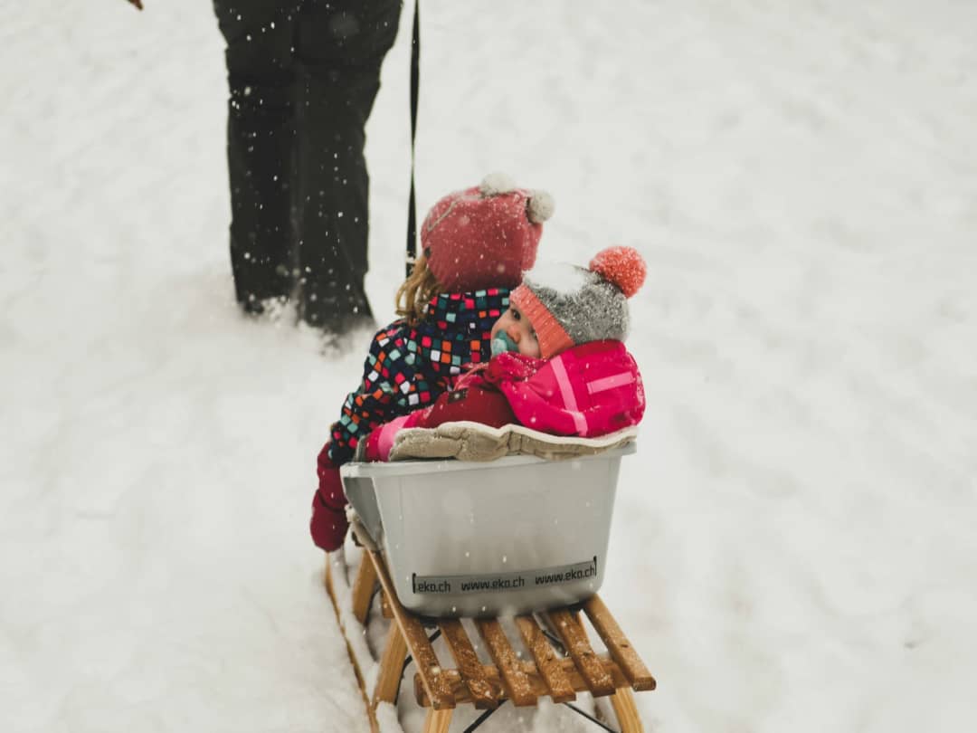 Two small children in woolly hats ride a sled pulled through falling snow in Interlaken in January