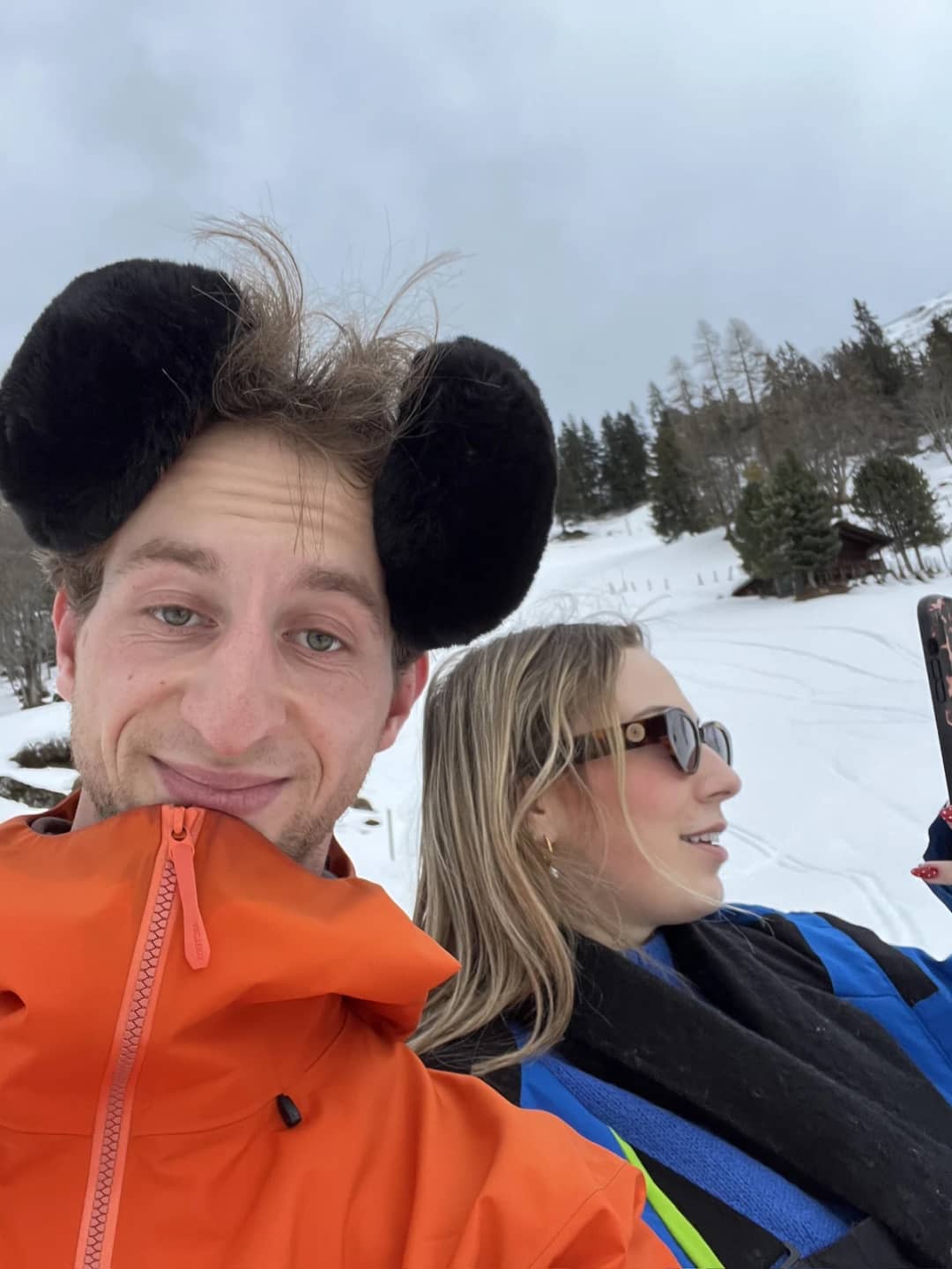 Pierre and a guest in sunglasses take a selfie on a snowy meadow above Isenfluh, pine trees and a chalet behind