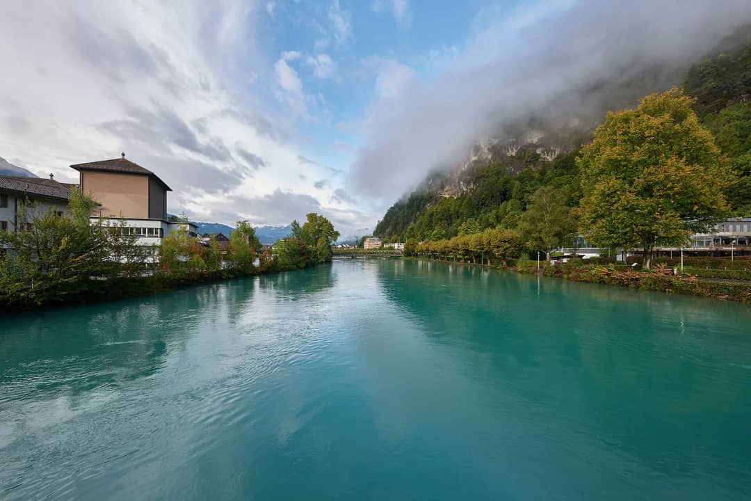 Turquoise Aare canal flowing through Interlaken, lined with trees and houses, a wooded slope in low cloud.