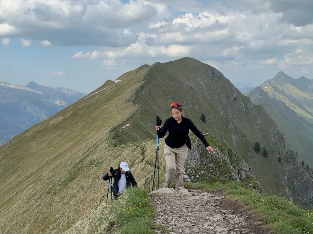 Two hikers with poles climb the grassy Augstmatthorn ridge path, patches of snow on the slopes under clouds.