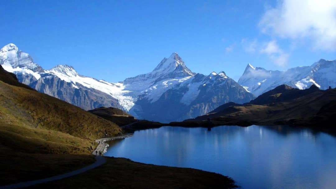 Calm Bachalpsee reflecting the snowy Schreckhorn, a few hikers on the lakeside path beneath a clear blue sky.