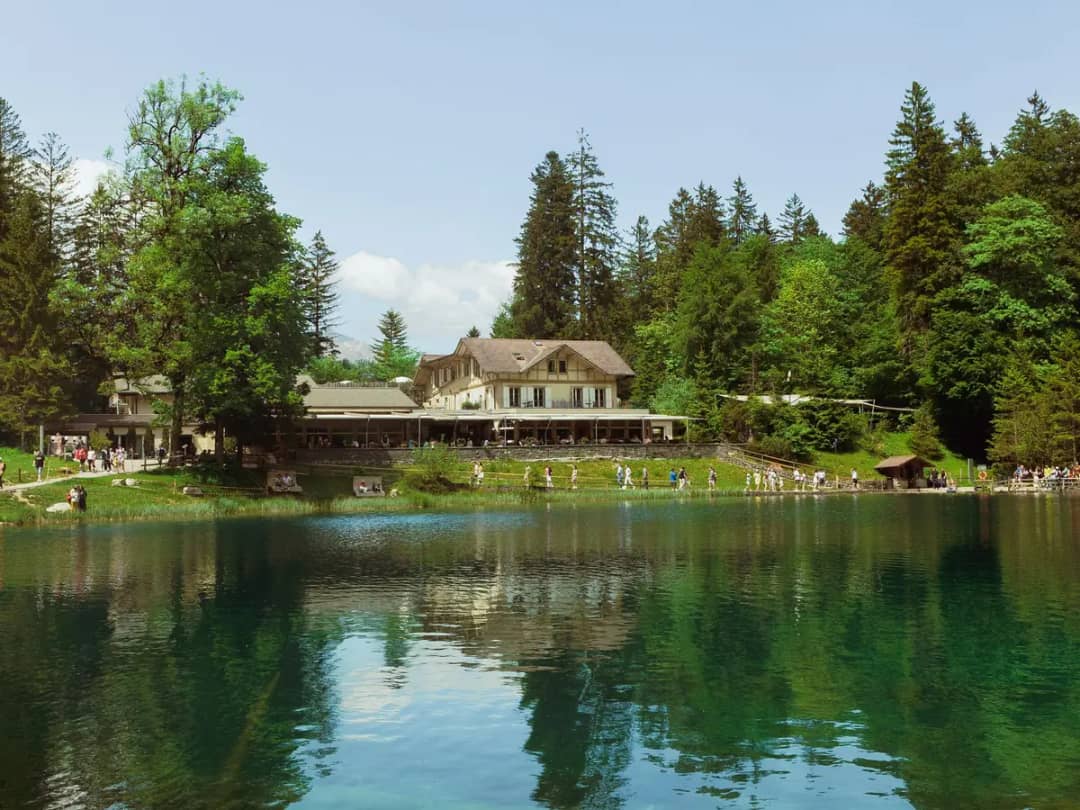 Hotelgebäude am Blausee und belebter Uferweg am grünen Wasser, hohe Fichten spiegeln sich im klaren Waldsee