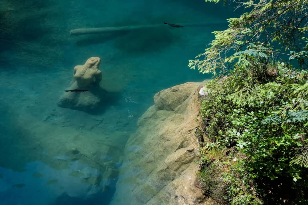 Statue de femme en pierre sous l'eau turquoise et limpide du Blausee, des truites nagent autour des rochers