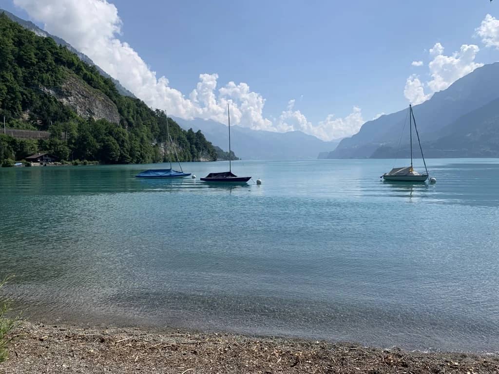 Drei vertäute Segelboote auf dem türkisfarbenen Brienzersee vor einem Kiesufer bei Bönigen, bewaldete Hänge