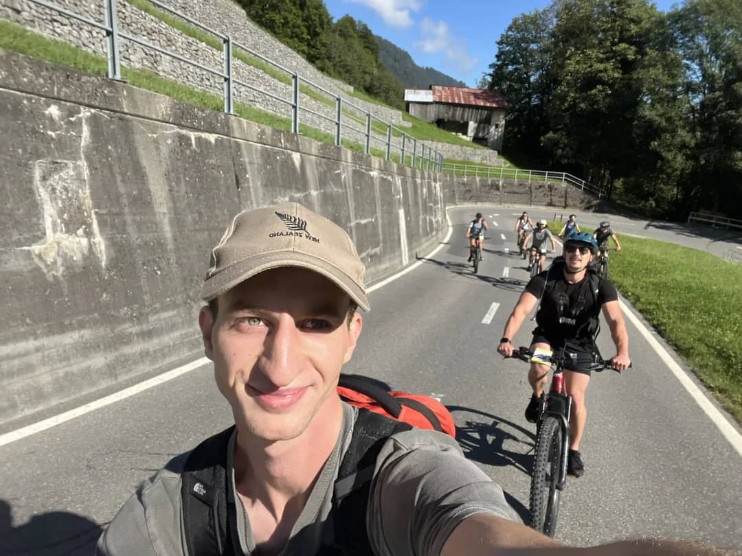 Lächelnder Guide mit Kappe macht ein Selfie, dahinter eine Velogruppe auf einer Talstrasse bei Interlaken
