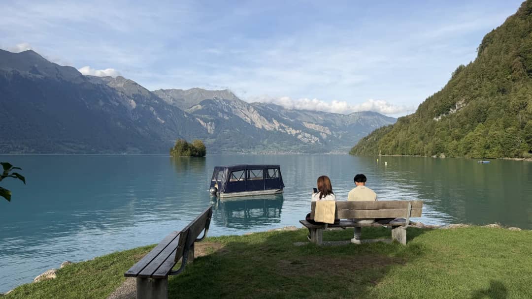 Pareja sentada en un banco de madera en Iseltwald frente a un barco y el islote del lago de Brienz, de tarde