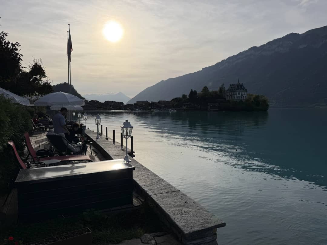 Seeterrasse mit Laternen in Iseltwald, dunstige Abendsonne über dem Brienzersee und kleines Schloss hinten