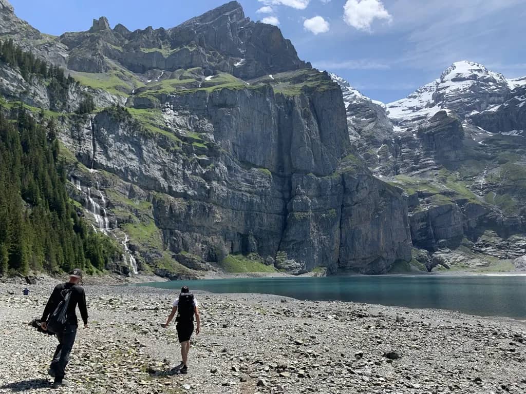 Dos senderistas cruzan la orilla pedregosa del Oeschinensee bajo acantilados grises y cumbres nevadas