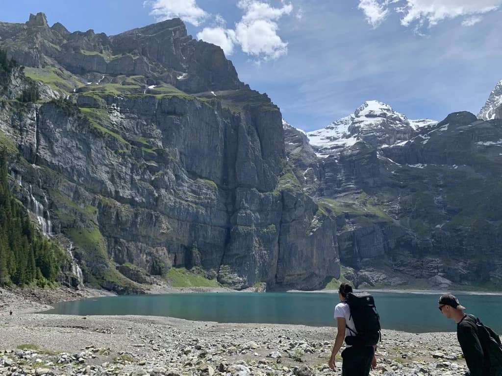 Dos senderistas con mochila en la orilla pedregosa del Oeschinensee, agua turquesa bajo acantilados nevados