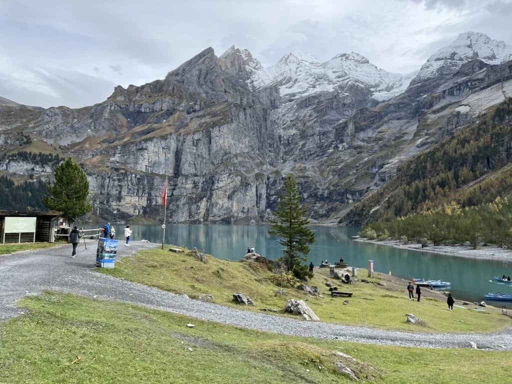 Visitantes en el camino de grava junto al Oeschinensee en octubre, barcas en el agua y nieve en las cumbres