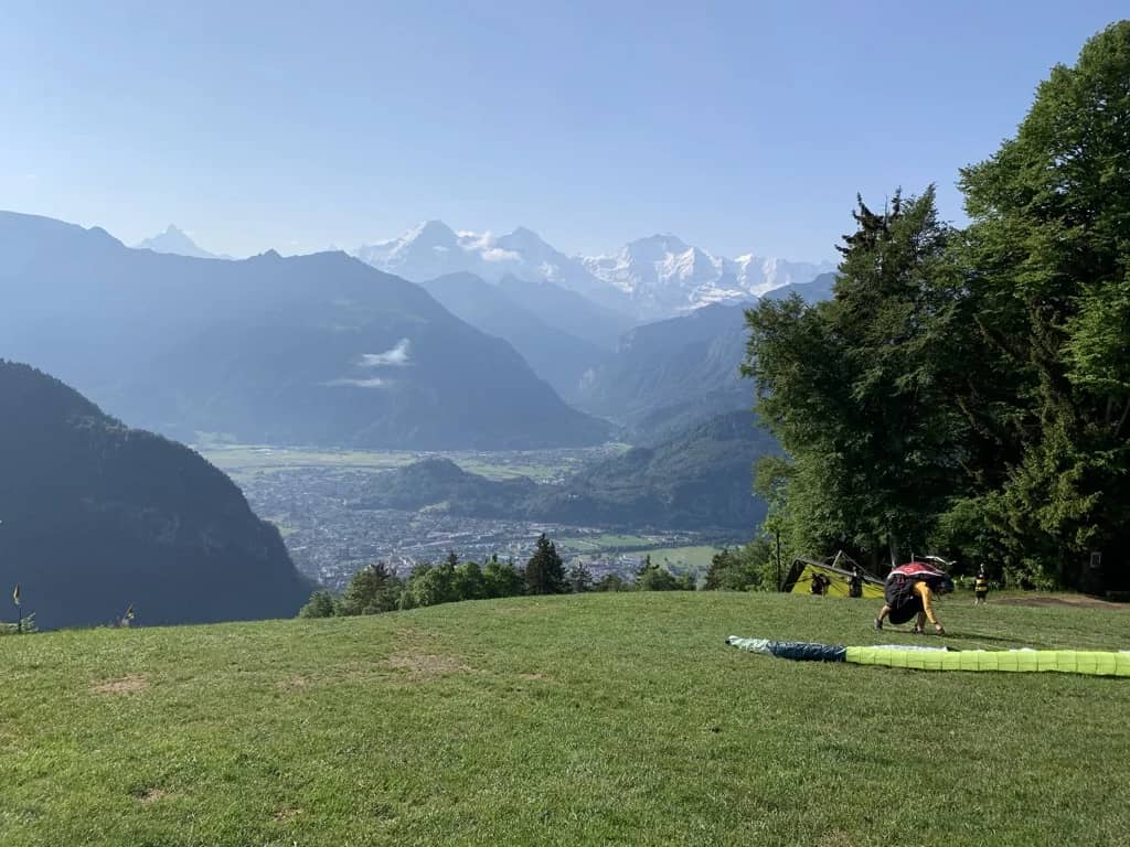 Pilot laying out a green canopy on the Beatenberg launch meadow above Interlaken, Eiger and Jungfrau in haze