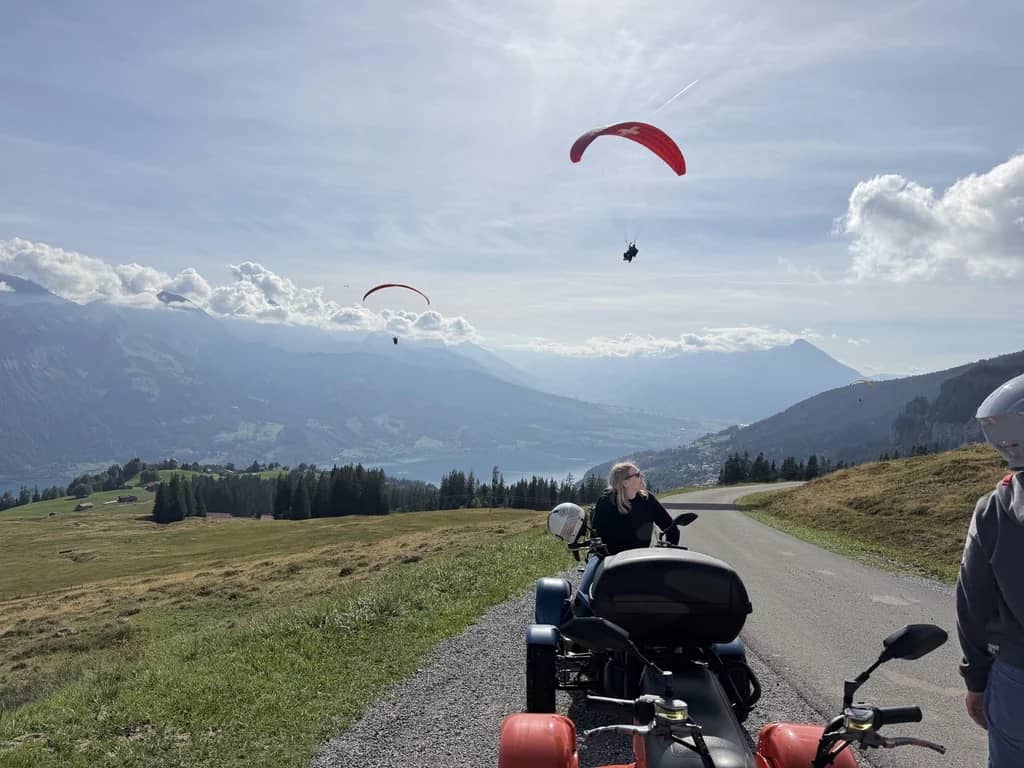 Two red paragliders circling above Lake Thun and the Niesen, seen from a road on the Beatenberg slopes