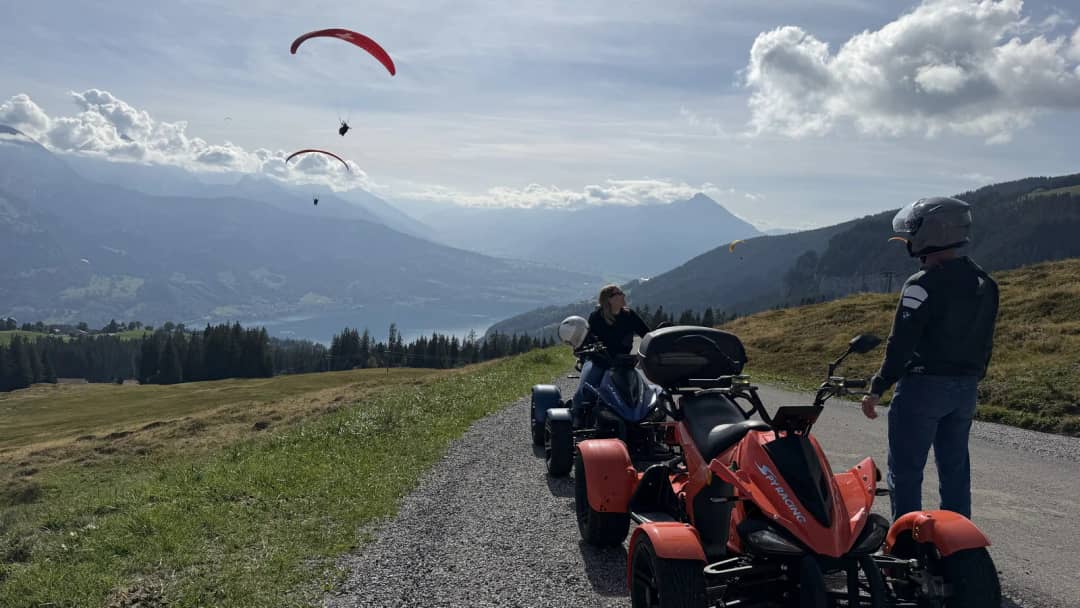 Two quad riders in helmets stop on a gravel track above Lake Thun as red paragliders circle the valley.