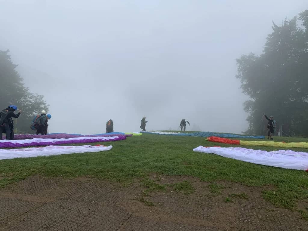 Pilots waiting beside canopies spread on the grass of a fog-covered launch site above Interlaken, zero view