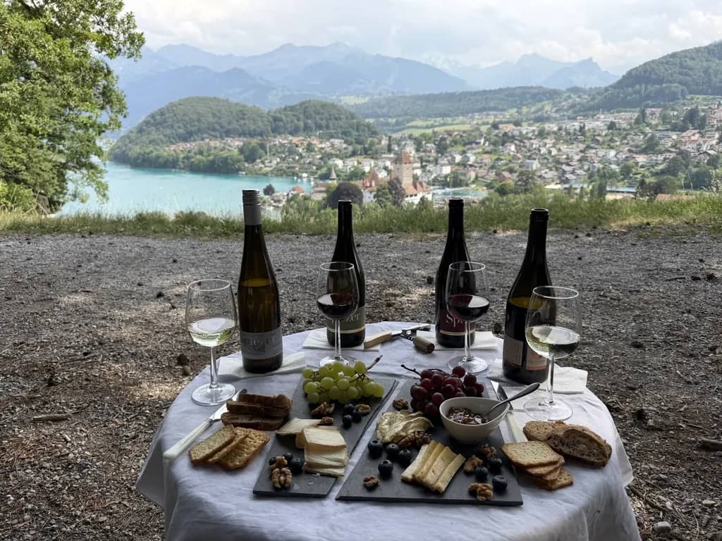 Mesa de cata con cuatro botellas de Spiez, copas, queso y uvas en una colina sobre la bahía del lago de Thun
