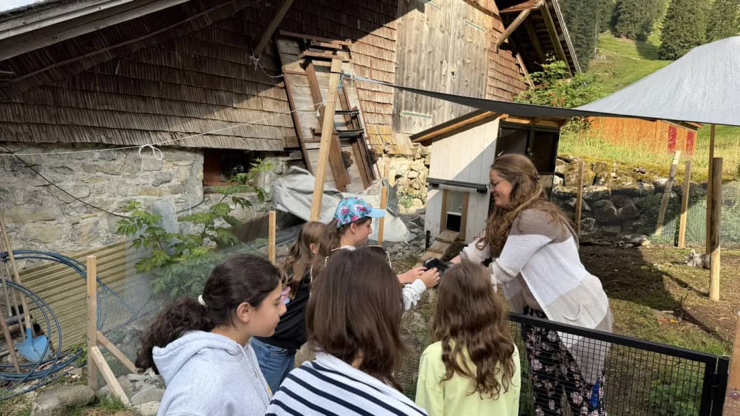 Daniela montre un petit animal à de jeunes clients du Farm Tour devant une ferme d'alpage en bois, Justistal