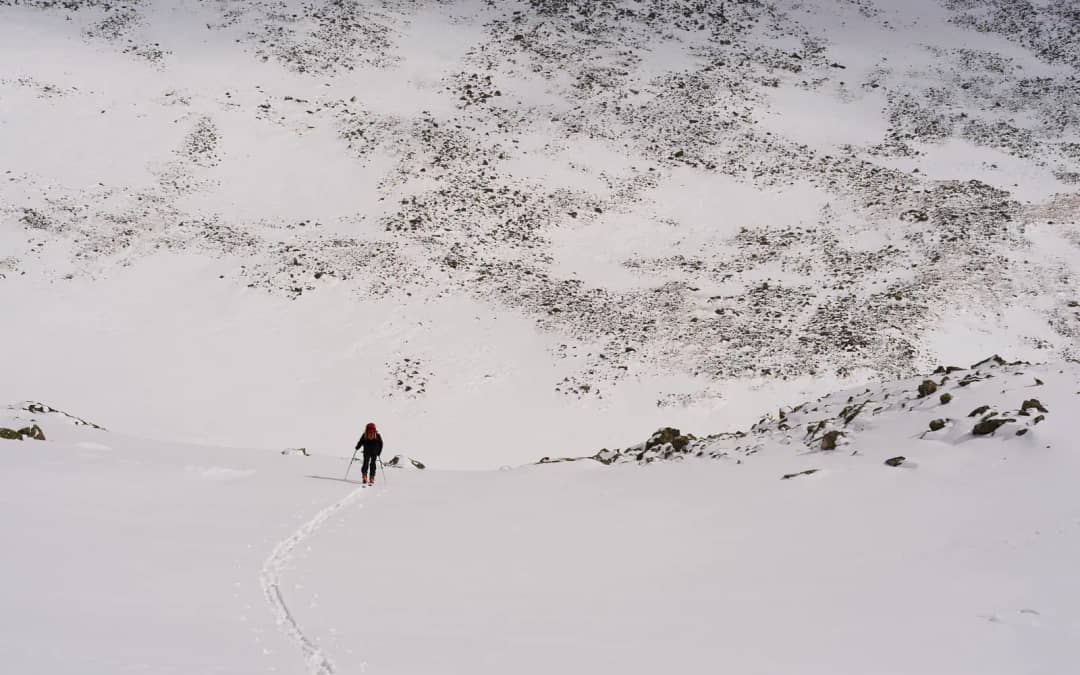 Lone walker crosses a wide snowfield below a rocky slope, open terrain where avalanche risk begins