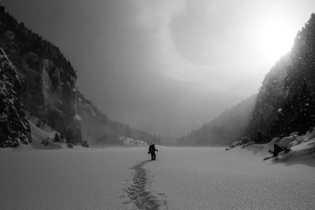 Lone hiker heads into a misty snowy valley between dark forests, footprints in fresh snow