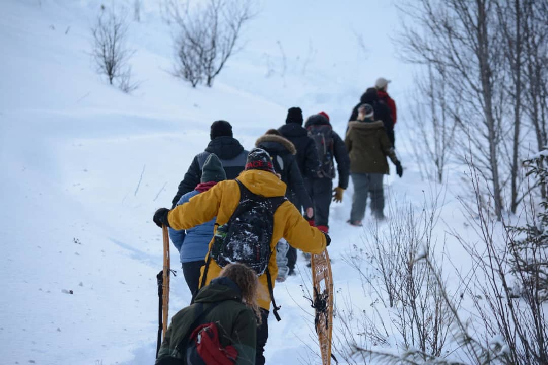 Group of snowshoers in winter jackets climbs a snowy slope in single file, some carrying wooden snowshoes