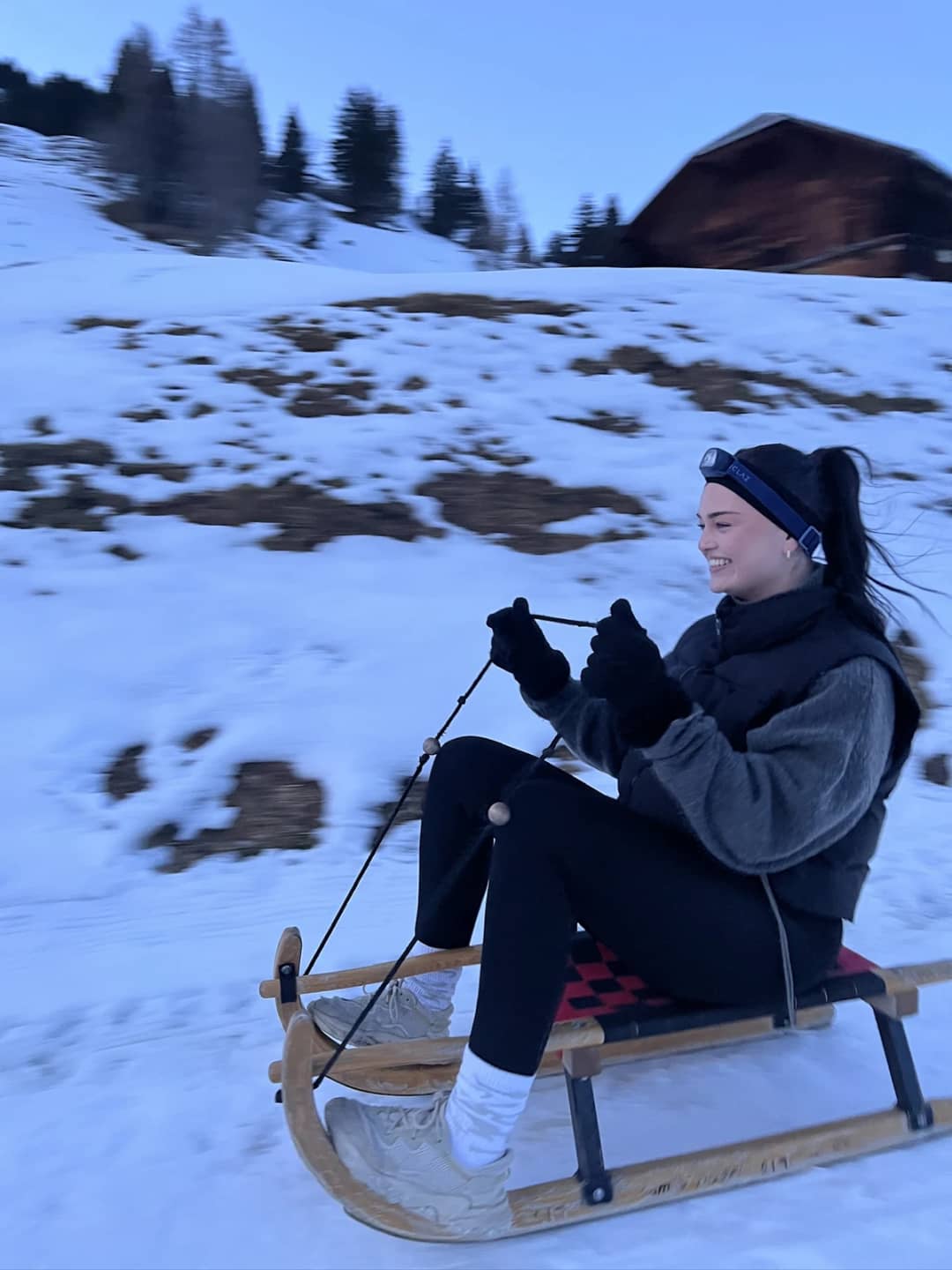 Young woman sledding down the snowy slope at Sulwald above Isenfluh, a chalet roof behind, on a January evening