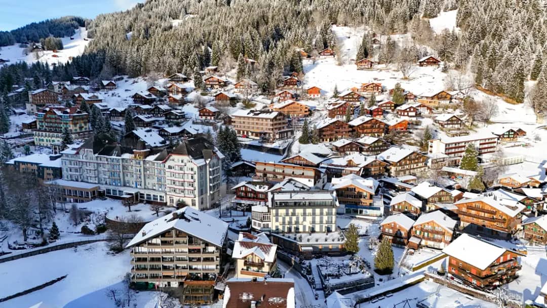 Vista aérea de Wengen, pueblo sin coches, con chalés y hoteles nevados bajo un bosque de invierno al sol