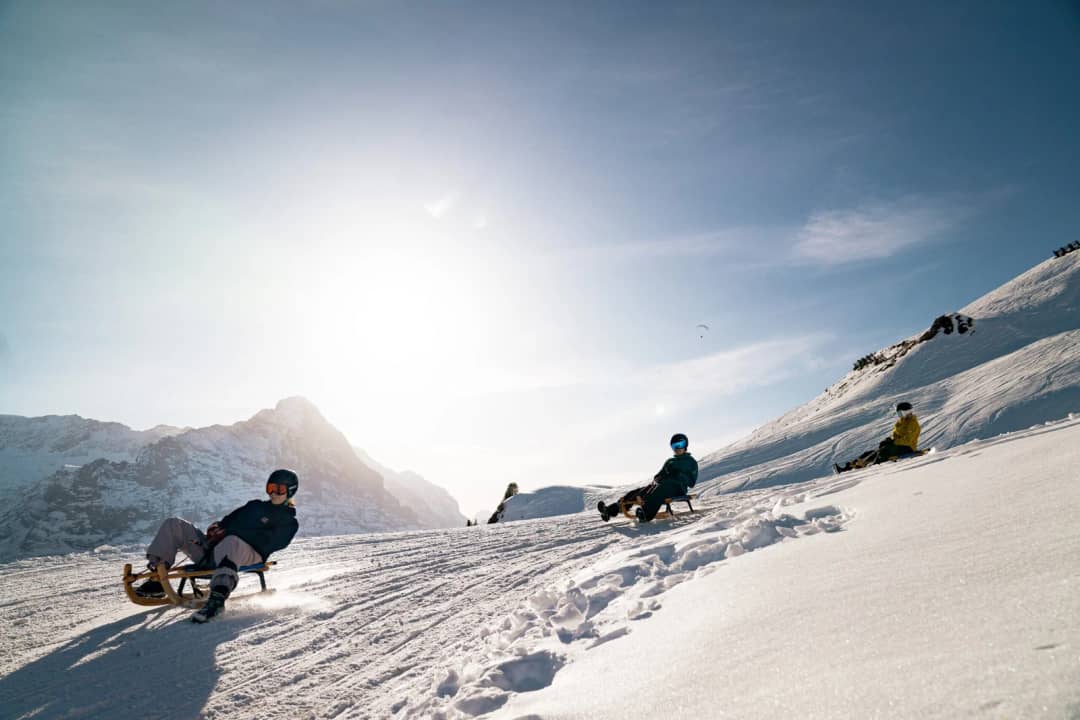 Persona en trineo inclinada en una curva nevada de la pista de First a Schreckfeld, con sol bajo tras las cumbres