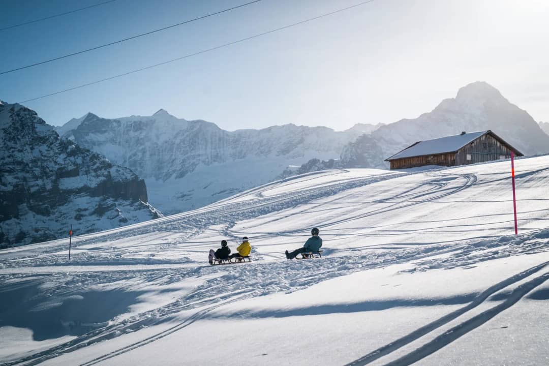 Personas en trineo junto a una cabaña de madera en la pista nevada de Grindelwald First, con cumbres detrás