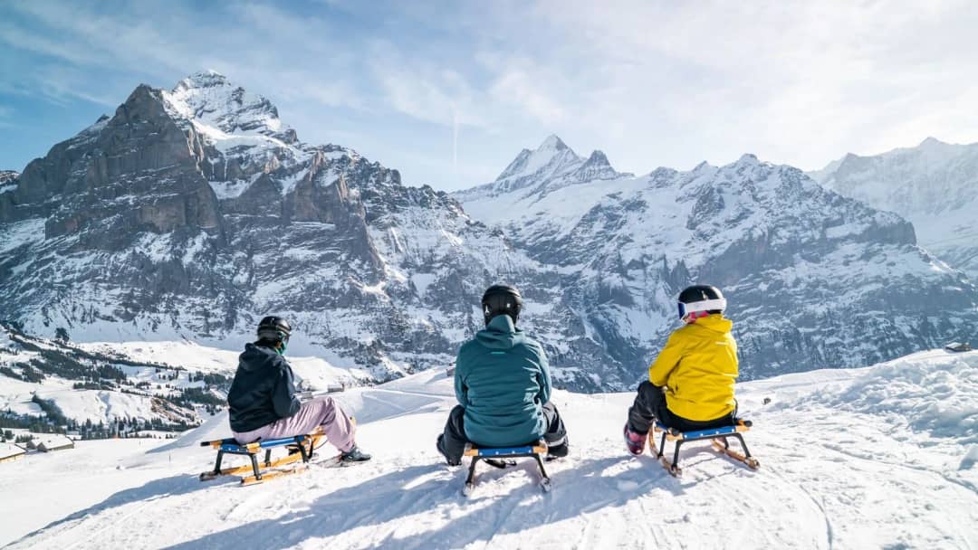 Tres personas descansan en trineos de madera sobre Grindelwald First, frente al Wetterhorn nevado, con sol