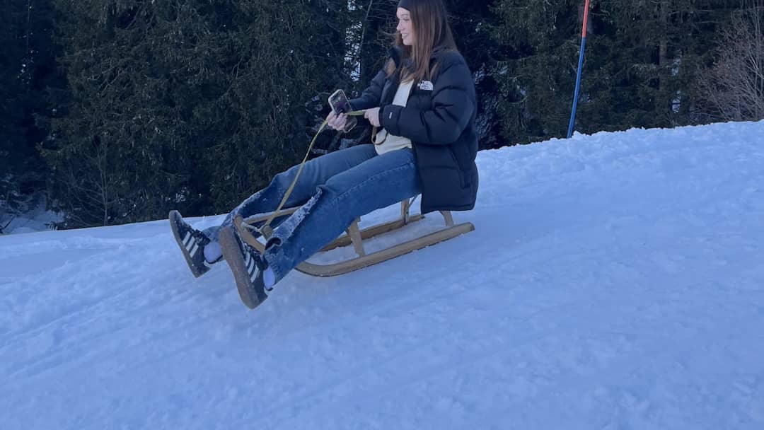 Young woman on a wooden sled, sitting upright with her feet forward, on a snowy forest sled run in Mürren