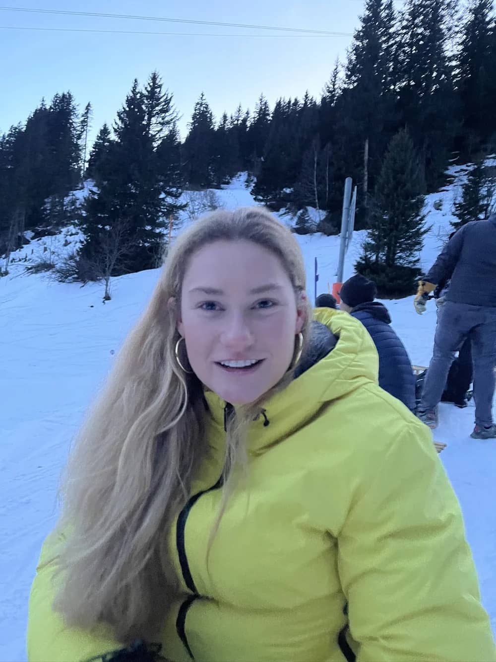 First-time sledder in a yellow ski jacket smiling on a snowy sled run above Mürren, other sledders behind her