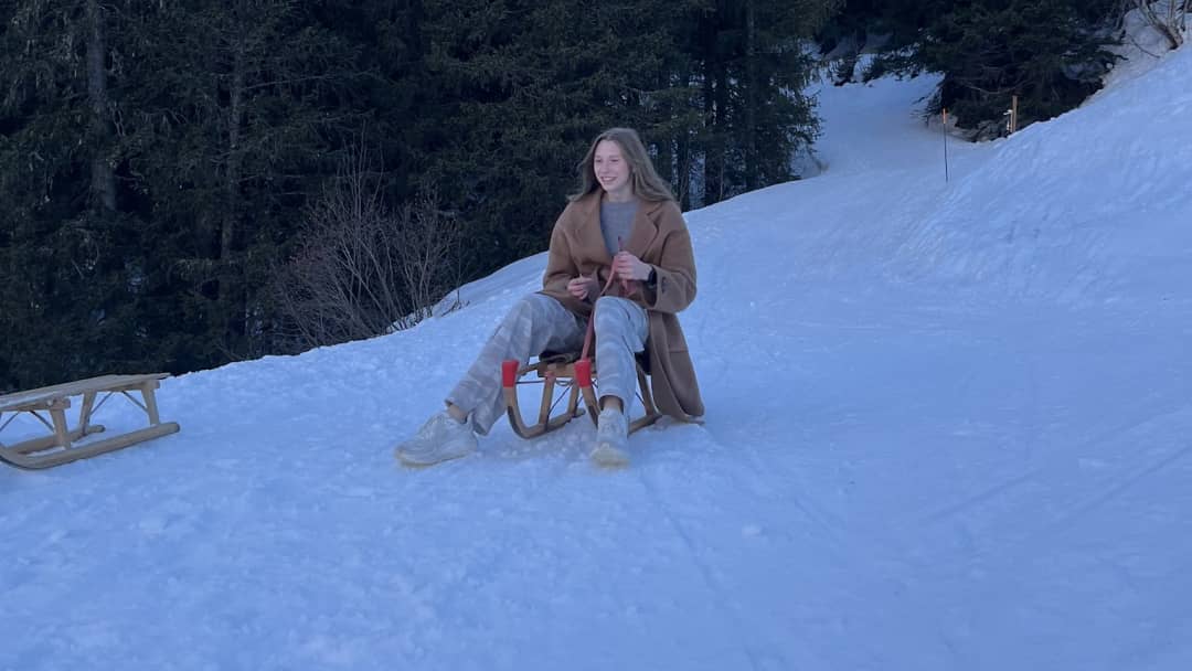 Jeune femme riant sur une luge en bois, piste forestière enneigée au-dessus de Mürren, une autre luge à côté
