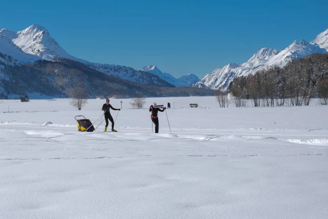 Two adults on cross-country skis pulling a child in a sled across a snowy Swiss valley in winter