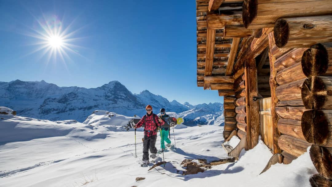 Two snowshoers pass a log hut in bright sunshine on a signposted snowshoe trail above Grindelwald