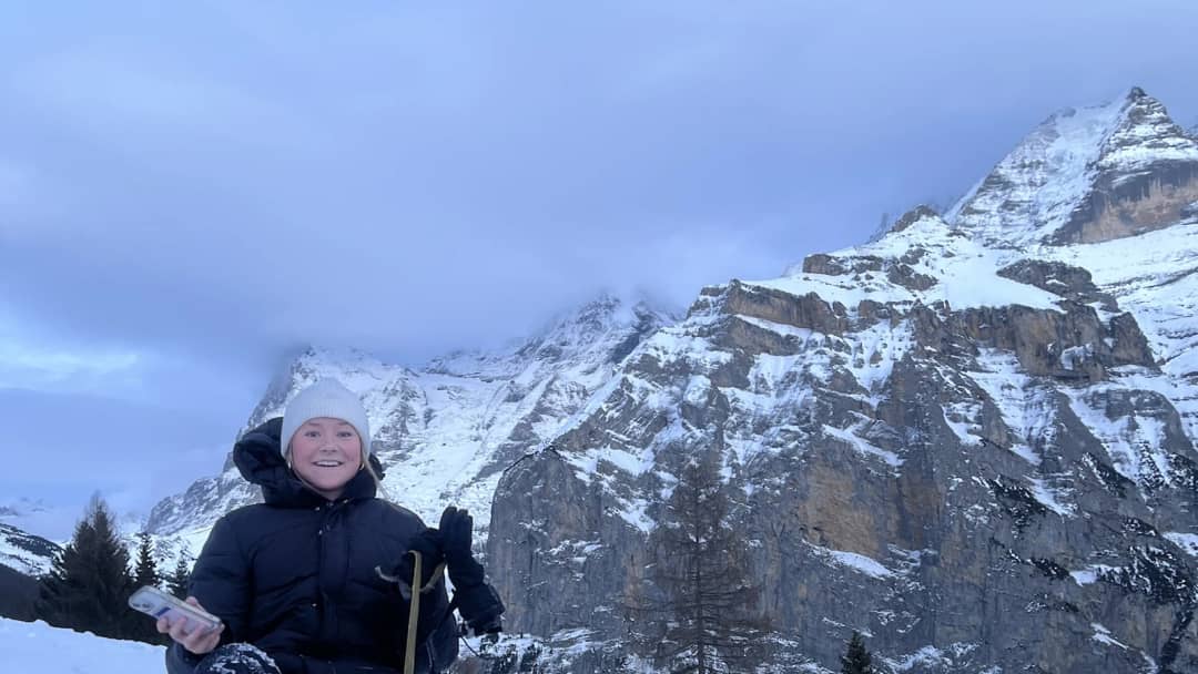 Gast in schwarzer Skijacke und weisser Mütze auf einem Holzschlitten unter den Felsen von Mürren am Abend